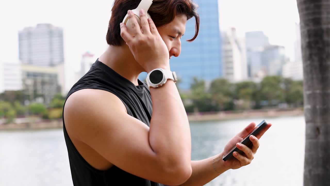 A fit Asian man wearing headphones and a smartwatch uses his smartphone in Benchakitti Park