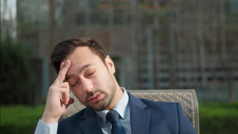 tired sad businessman in suit massaging head forehead and close eyes feeling pain or stress while sitting in city park - portrait close-up slow motion