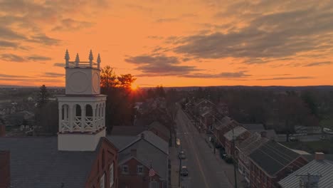 a drone pull back view of a small town and a steeple at sunrise as it gets ready to break the horizon, with orange and reds on a spring sunrise with partial cloudy skies