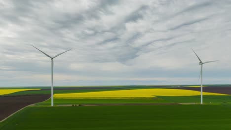 Aerial-hyperlapse-with-wind-turbines-on-a-green-field-and-yellow-rapeseed-on-a-cloudy-day