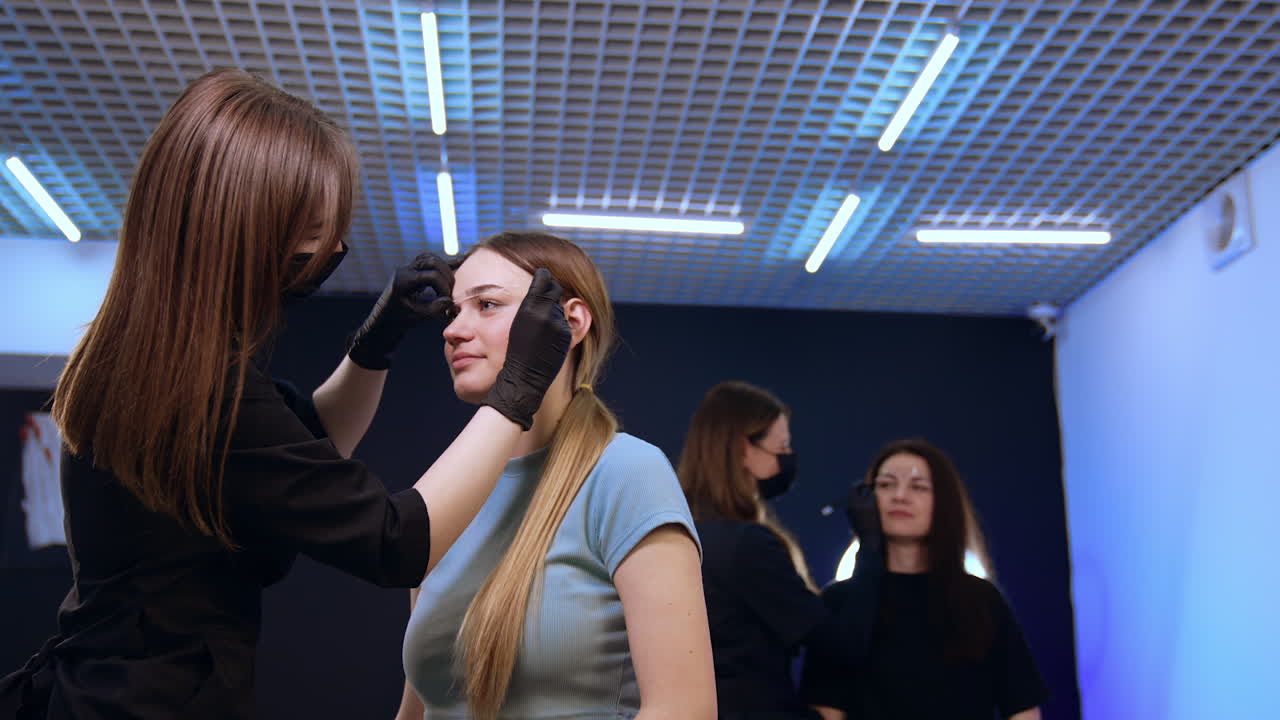 Brow master marking the lines of girl's eyebrows with the thread. Beautician draws lines with white pen to a woman at backdrop.