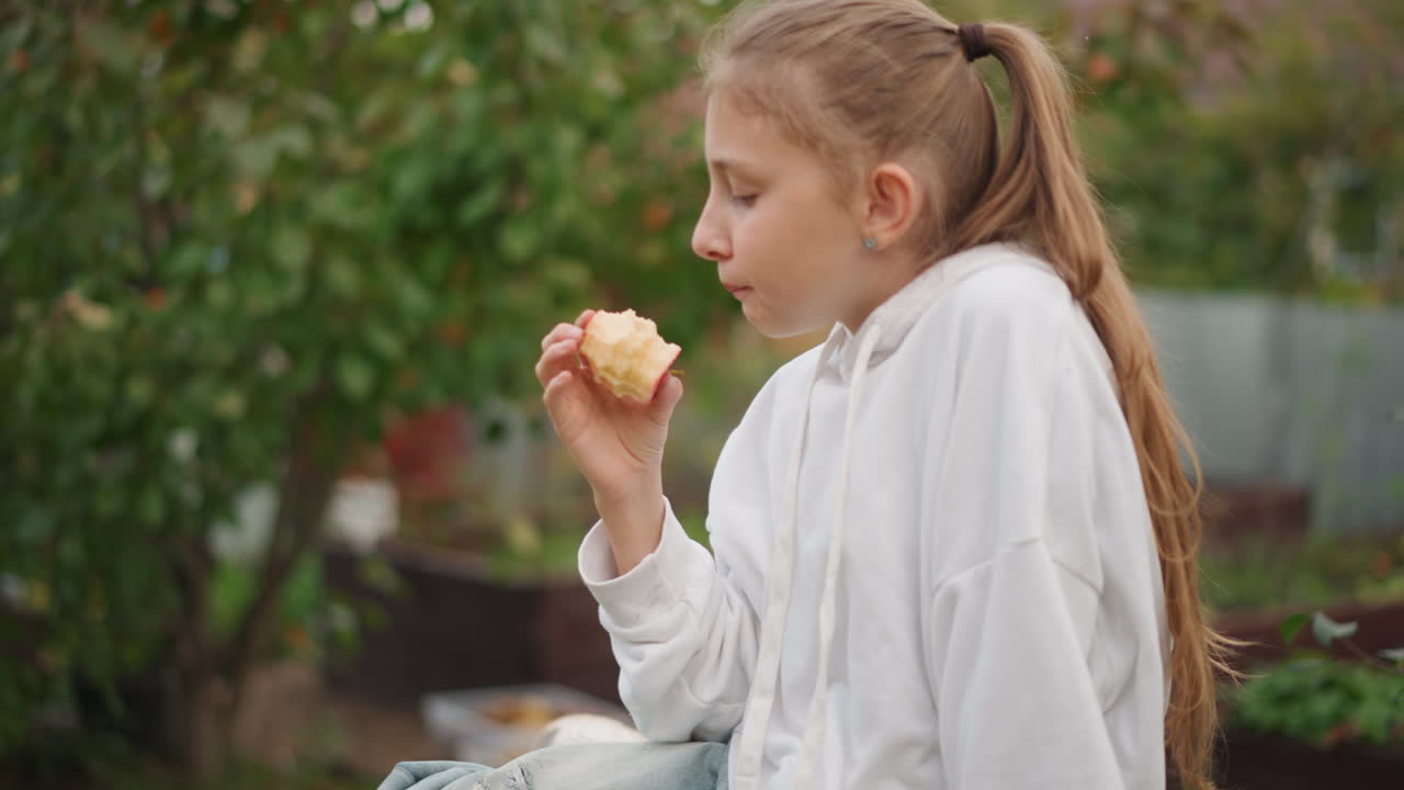 Young caucasian girl sitting on bench eating apple in backyard garden, white hoodie, long ponytail, relaxed pose on wooden plank seat, soft autumn light, greenery bokeh, slow thoughtful chew, candid