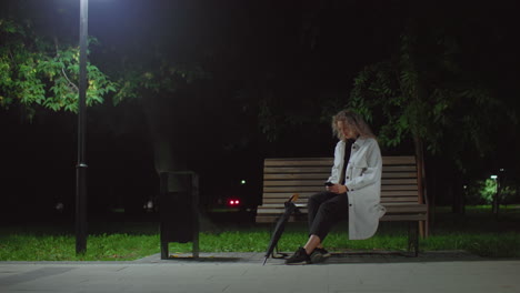 girl seated alone on park bench at night under glowing streetlamp, operating phone and gazing into distant space with wind swaying green leaves and red light glowing softly in dark background