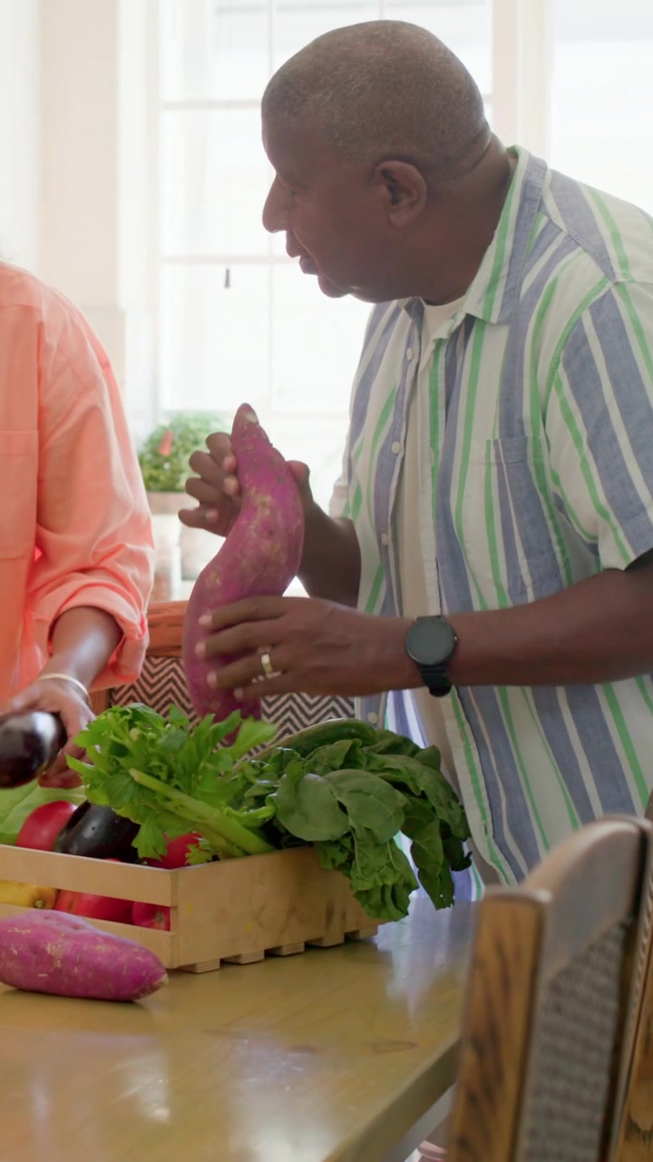 Vertical video: Entering kitchen with crate, man in striped sorting produce with wife prepping meal