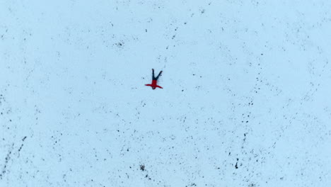 Man-With-Red-Jacket-Lying-on-His-Back-in-the-Snow,-Spiraling-Ascending-Aerial-Top-Down-Shot