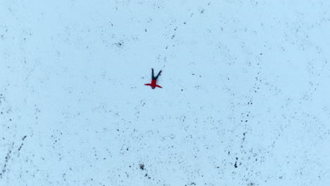 man with red jacket lying on his back in the snow, spiraling ascending aerial top down shot