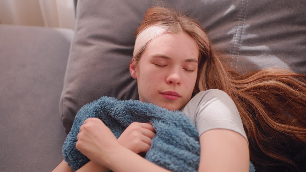 Close up top down view of dreamy woman resting on grey couch with soft blanket pulled to chin as sunlight reflects on her peaceful face while long hair flows across cushion in quiet warm setting