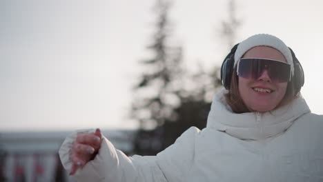 closeup of woman spinning with fluid choreography and radiant expression, wearing white puffer jacket, beanie, goggles and headphones as she dances on snow dusted urban plaza under winter sunlight
