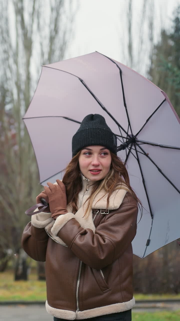 Signora allegra che tiene un ombrello mentre cammina nel parco in una fredda giornata nuvolosa, indossa un berretto di maglia nero e una giacca marrone in shearling, circondata da pini ed edifici urbani con una leggera nevicata sullo sfondo