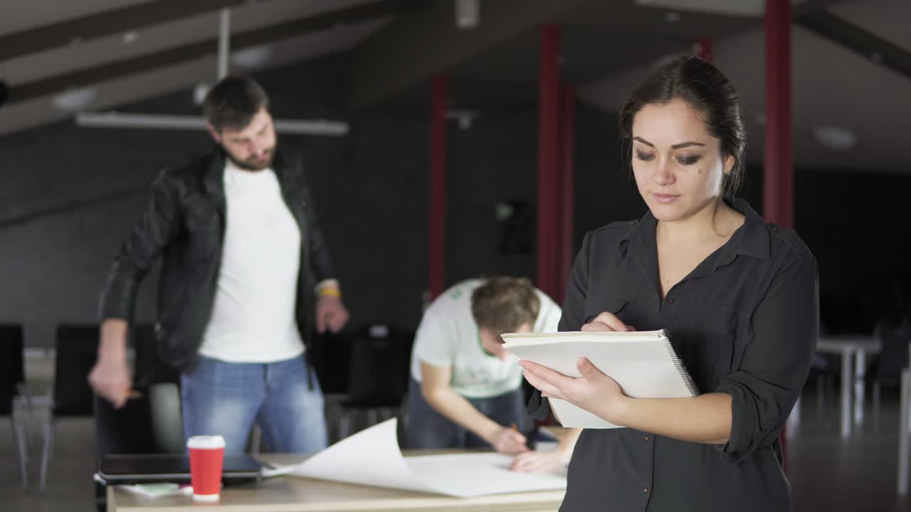 Portrait of a fashionable young professional woman holding folders with papers and taking notes in a stylish contemporary office. Office workers on the background. Shot in 4k