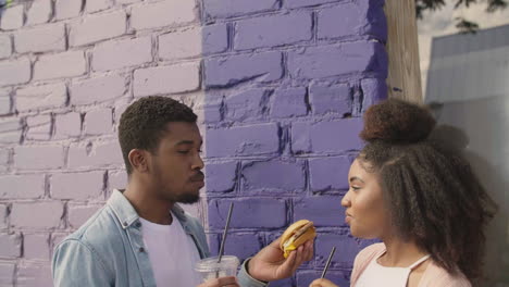 young couple sharing a tasty hamburger and drinking a cold drink together, while laughing leaning against a colourful brick wall