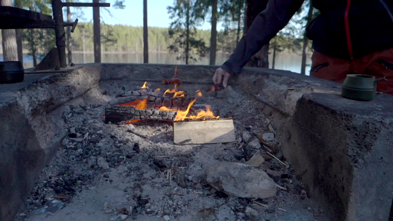 un excursionista asando un perrito caliente en una fogata frente a un pequeño lago