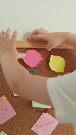 cute blond preschooler plays with paper and cork board slow motion. little boy presses cut pink figure trying to attach and lowers hand backside view closeup