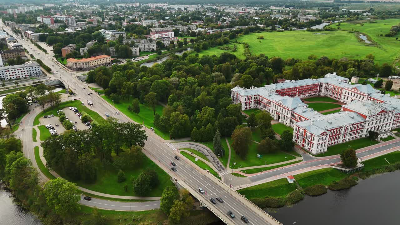 As the drone pulls away over Jelgava, the Baroque symmetry of Jelgava Palace unfolds beside the Lielupe River, with the bridge, parks, and city blocks receding into a layered urban panorama