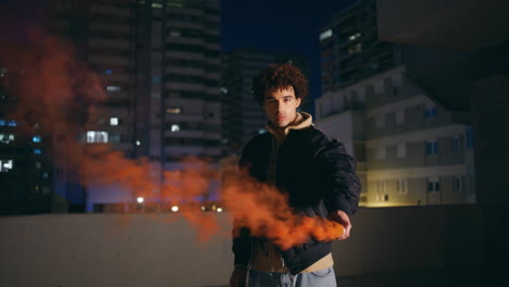 hipster holding smoke bomb at night city. curly guy teenager burning red grenade