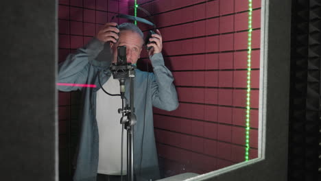 a man in a blue shirt and white inner shirt enters a red soundproof room, approaches a microphone, and takes a pair of headphones as he prepares to wear them, getting ready for a recording session