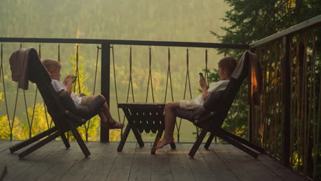 small kids play games on smartphones on transparent terrace at sunset. brother and sister rest in comfortable armchairs enjoying incredible view of dense forest