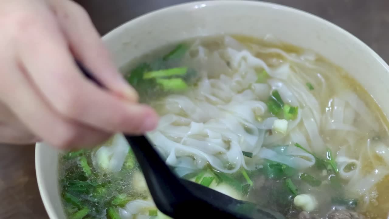 Close-up of a hand using a black spoon to enjoy noodle soup garnished with fresh herbs and greens.