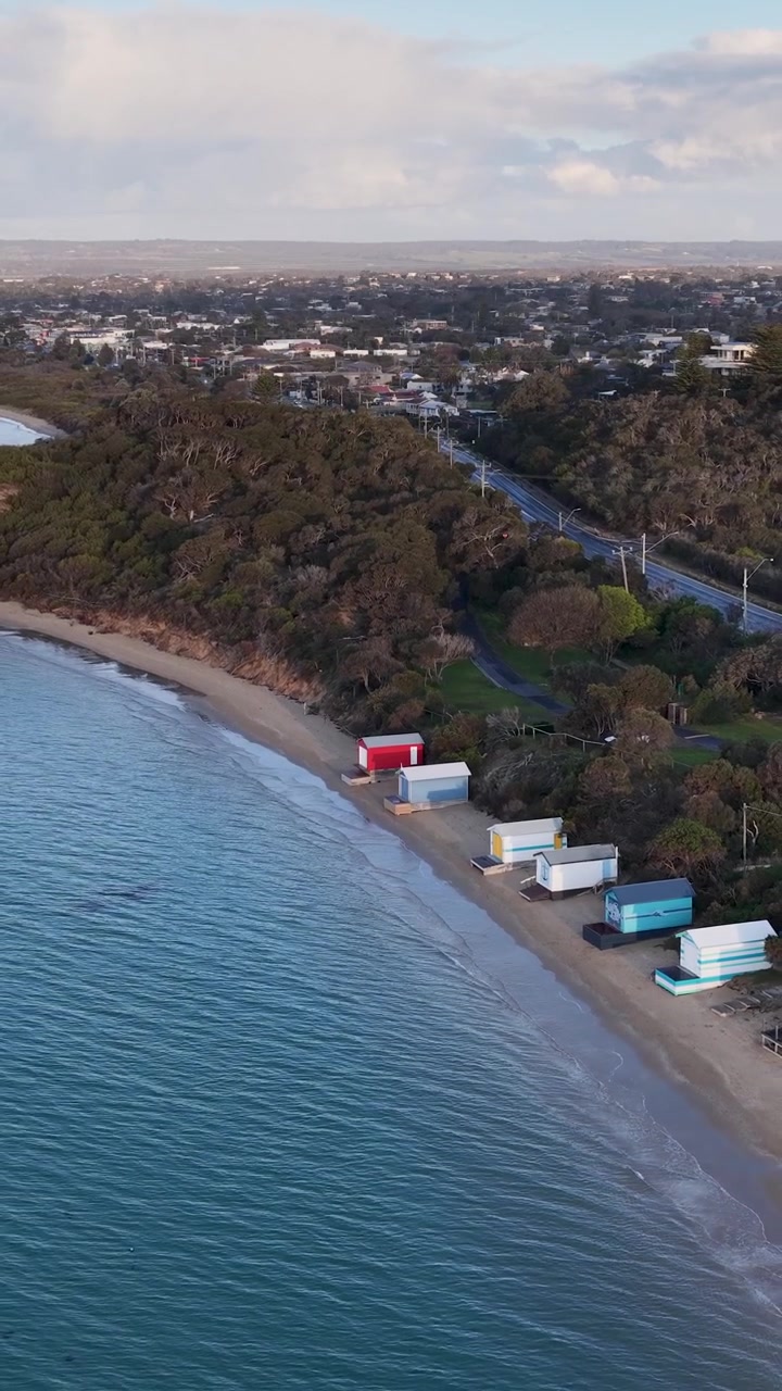 Un drone survole une plage de sable, des cabines de plage colorées, une mer calme et la végétation côtière au coucher du soleil.