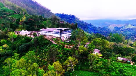 a sri lankan hillside features a prominent building nestled among green tea plantations, scattered rural homes, tropical vegetation, and layered mountain views under a cloudy sky.