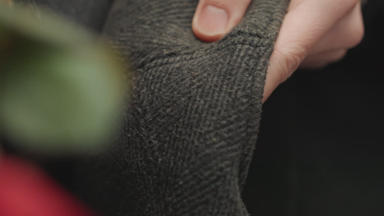 Gros plan de mains caucasiennes inspectant une casquette en laine, tissu tweed texturé et bouton supérieur dans le cadre, légère pression sur la couture, contrôle qualité tactile minutieux par le créateur dans un studio confortable, lumière naturelle douce, tamisée