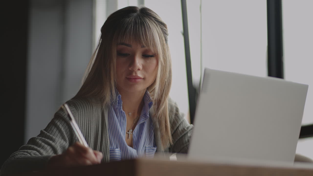 Asian woman working on her laptop and writing in his notebook sitting at a table. Working in coffee shop. woman looking to a laptop screen and making notes in her notebook. studying online.