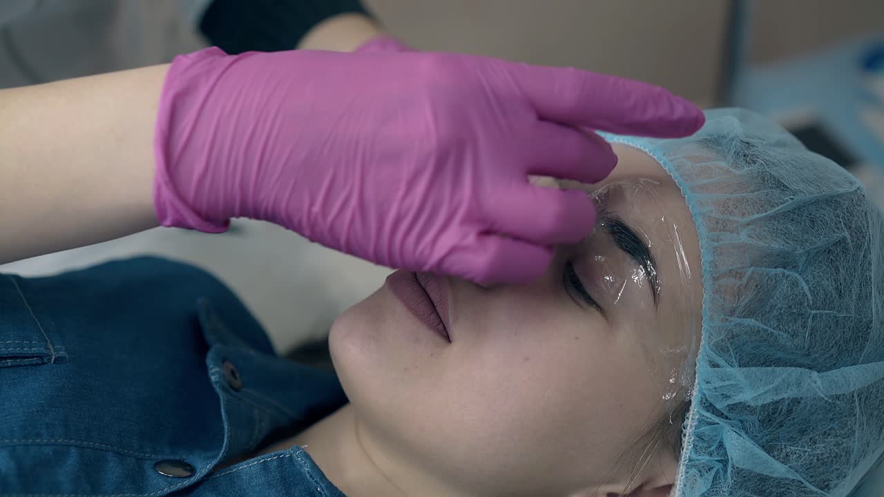 el cosmetólogo pone hojas de papel transparente en las cejas de la chica.