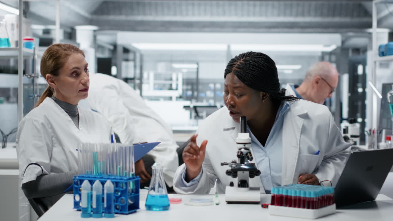 Biologist analyzes samples on microscope while coworker writes research notes
