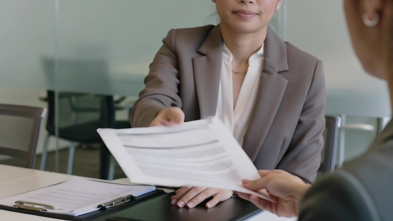 Donna asiatica in blazer grigio che consegna un foglio stampato attraverso il tavolo della riunione per la revisione di un collega.