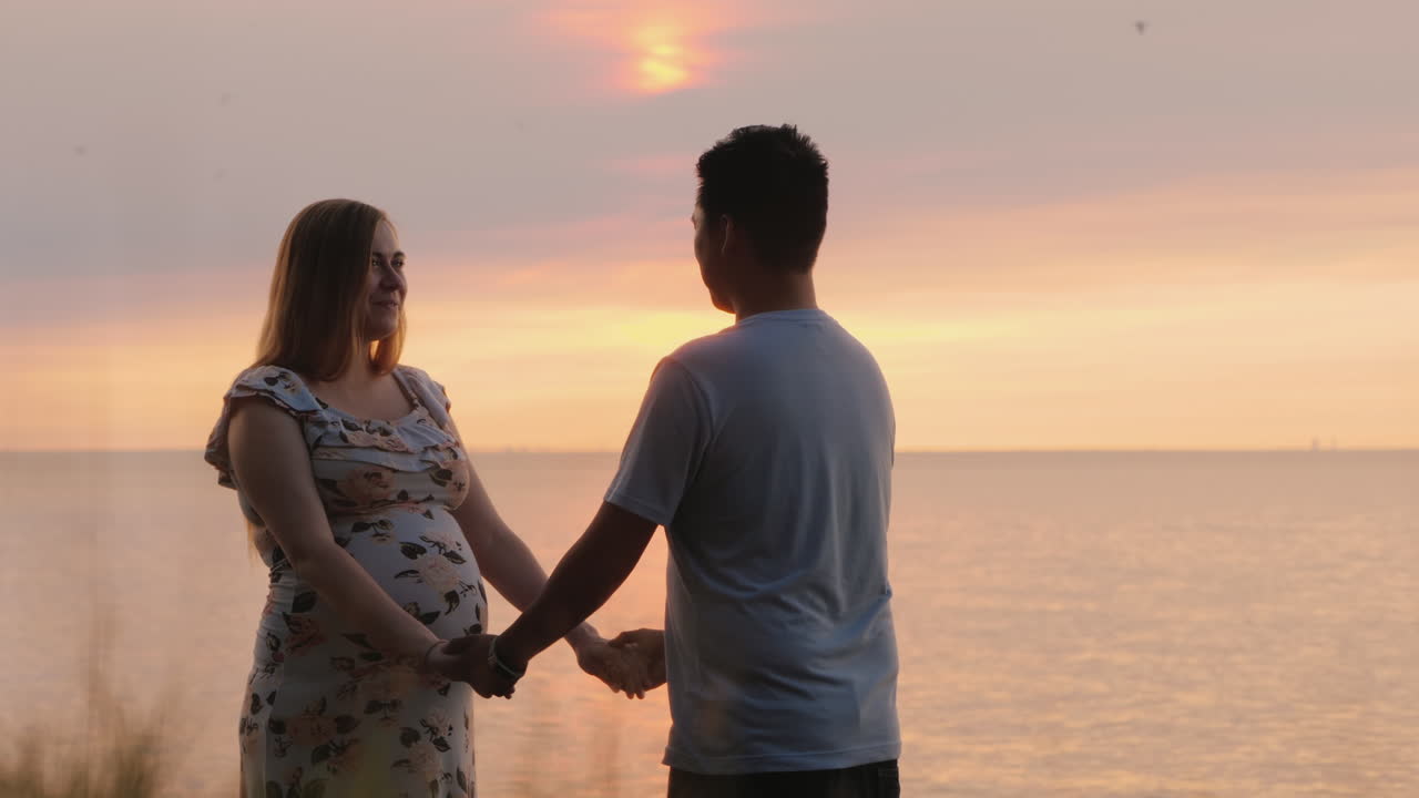다민족 몰트 커플이 바다 위의 일몰을 배경으로 손을 잡고 있다.