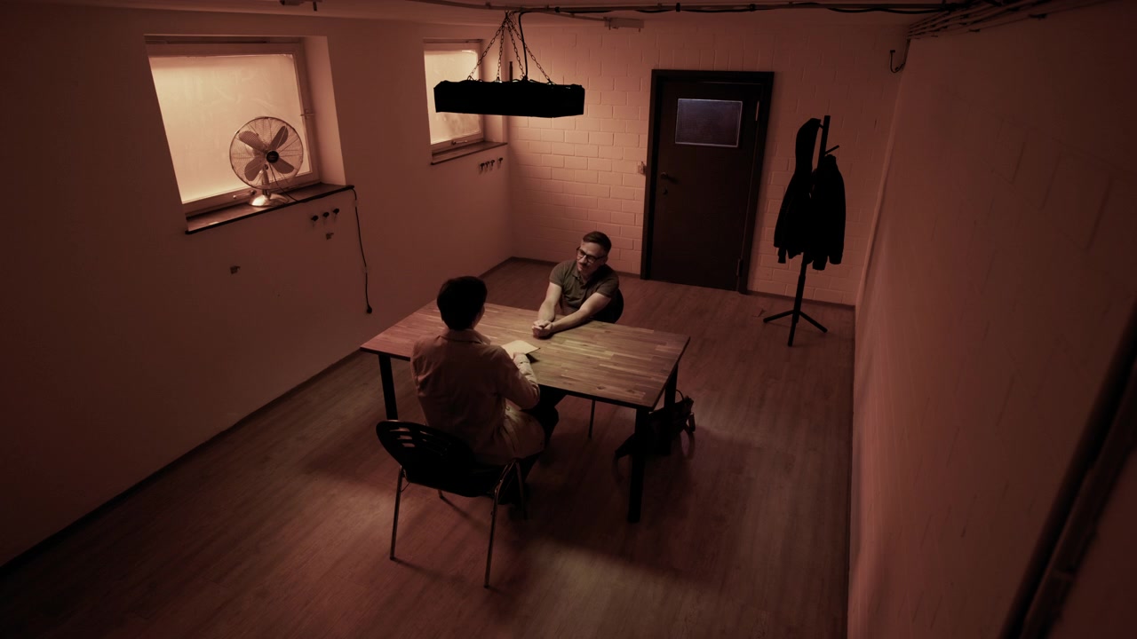 Scena cinematografica in una sala interrogatori con un detective che interroga un sospettato attraverso un tavolo. Temi di illuminazione suggestiva, tensione, indagine, crimine, mistero e narrazione drammatica