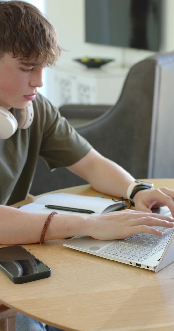 Vertical video: Male teen typing on silver laptop at home, checking notebook, wearing white headset