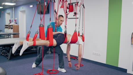 client male is undergoing rehabilitative physiotherapy on a special suspension rope system. young man is doing workout on a new equipment ropes with the help of the trainer. modern rehabilitation center.