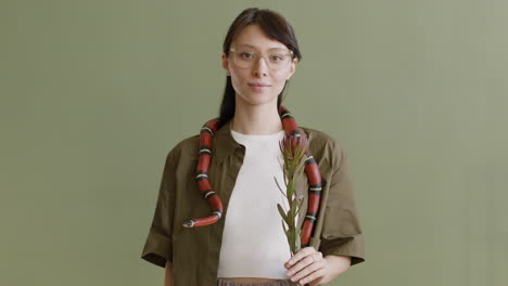 portrait of a young woman with a pet snake around her neck holding a twig and looking at the camera on a green background