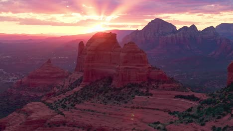 Incredible-sun-rays-pierce-clouds-spreading-pink-glow-above-Merry-Go-Round-red-rock-Sedona-Arizona,-drone-landscape