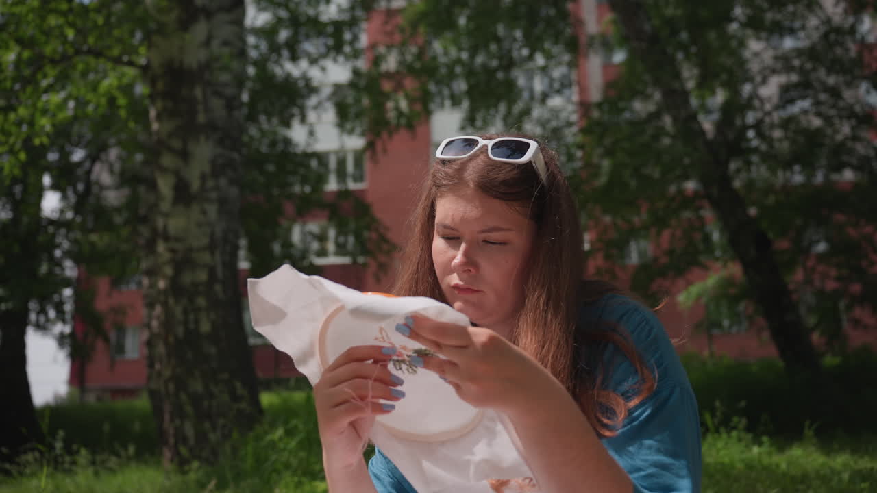 Young woman sitting on grass during sunny day, focusing carefully while embroidering colorful pattern on fabric hoop, expressing creativity in peaceful outdoor environment surrounded by green trees