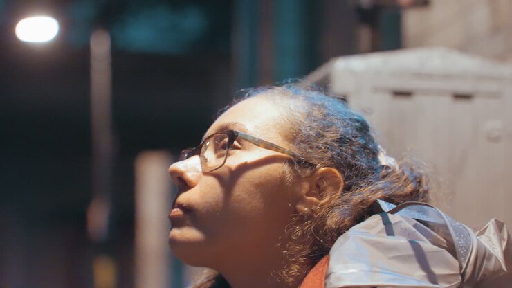Late night commuter young woman waiting at bus shelter holding snack, hair tied in bun, reflective jacket catching lamplight, empty platform and patient gaze, cinematic city atmosphere