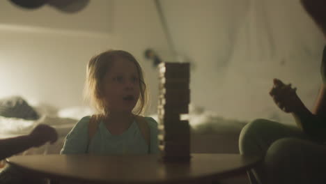 young woman starts game by pulling wooden block from tower and explaining rules to children. family spends game night sitting at small cozy table