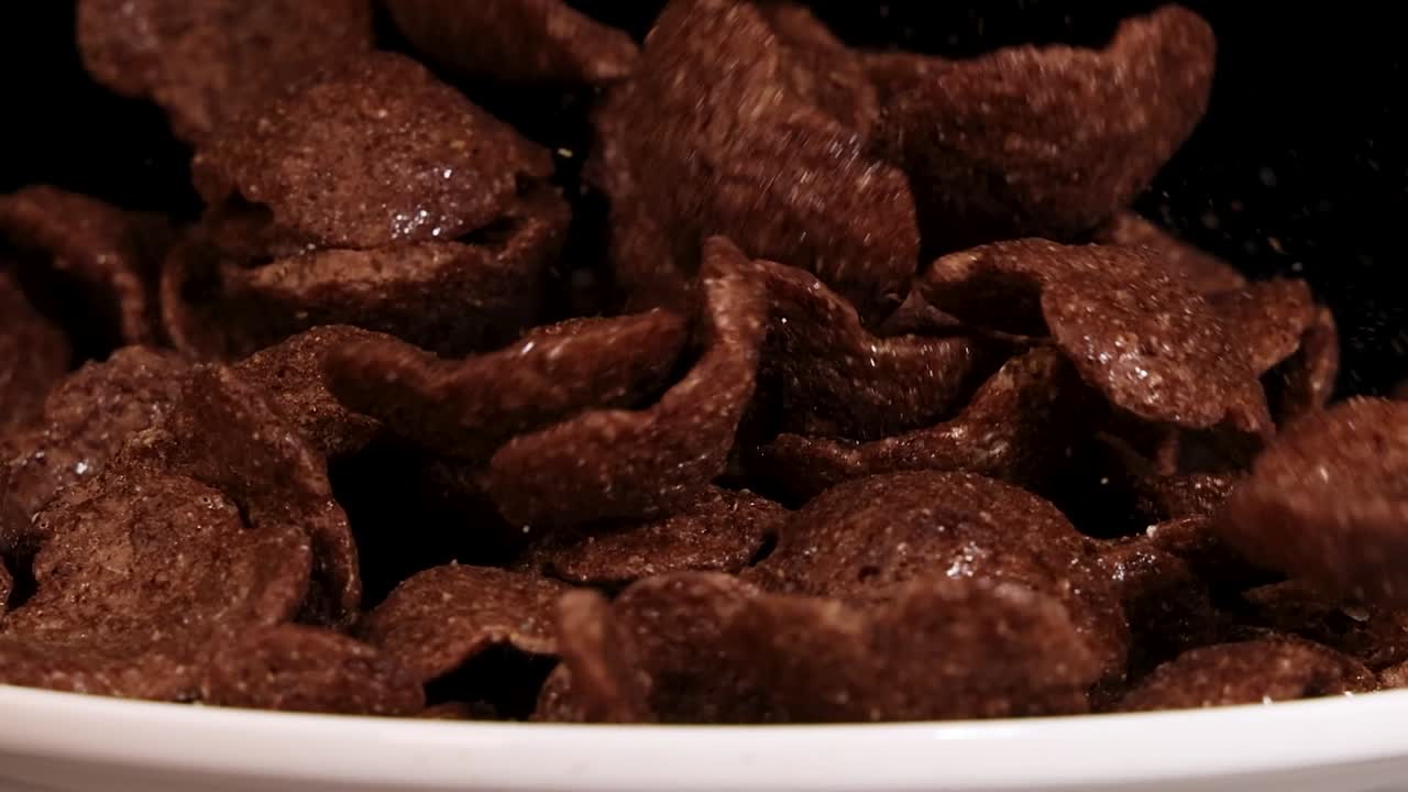 Chocolate cereal flakes cascade into a white ceramic bowl against a dark black background