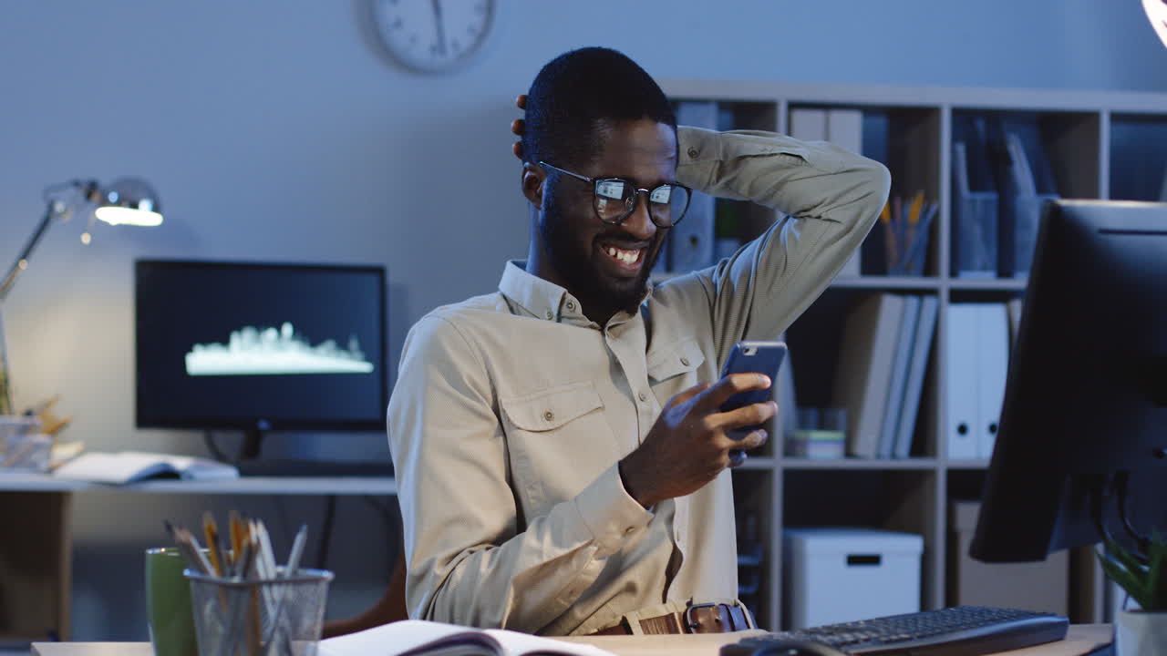 Smiled Man Sitting At Desk Chatting On His Smartphone While Resting A Minute In The Office At Night