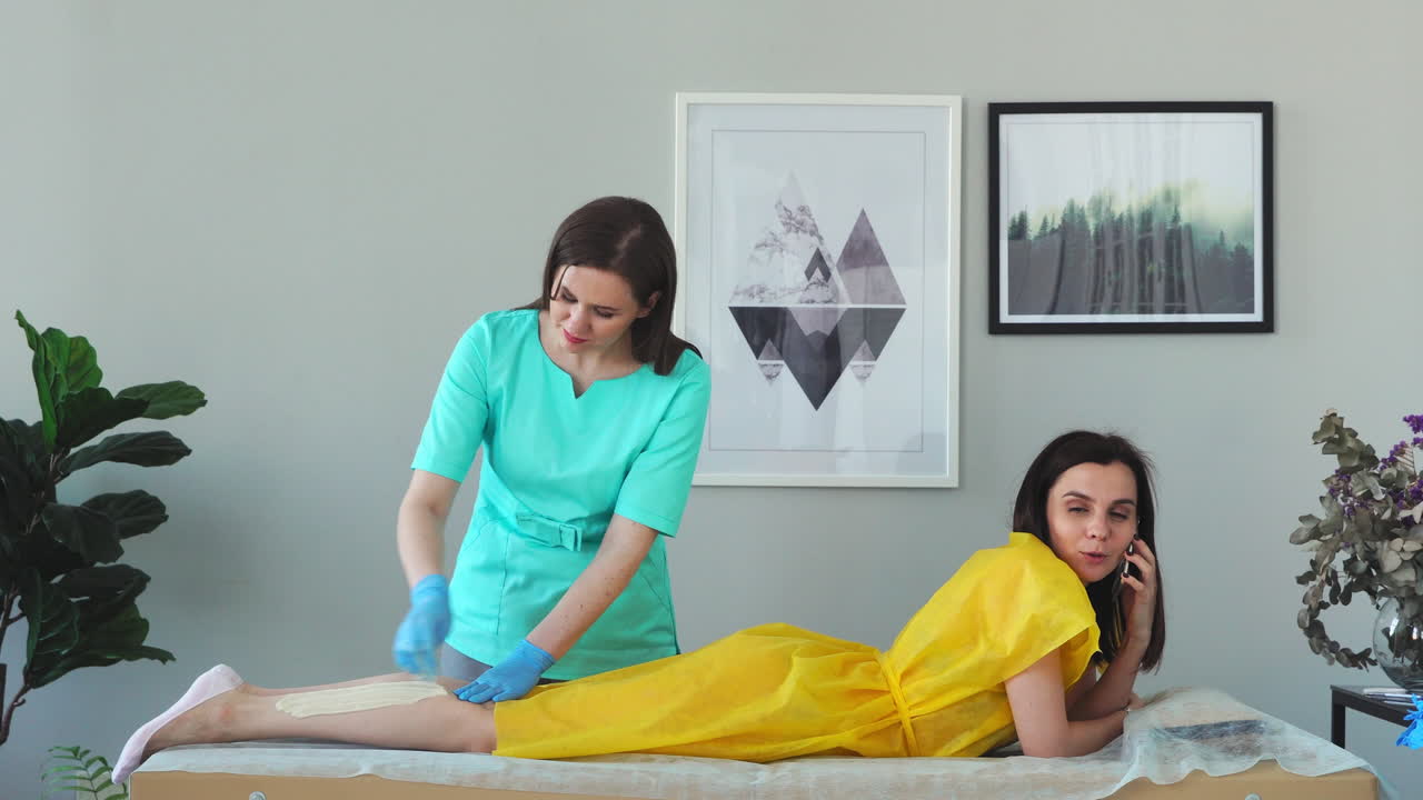 девушка разговаривает по телефону и улыбается во время удаления волос на ногах в салоне красоты. удаление волос на ногах сахаром. мастер удаления волос