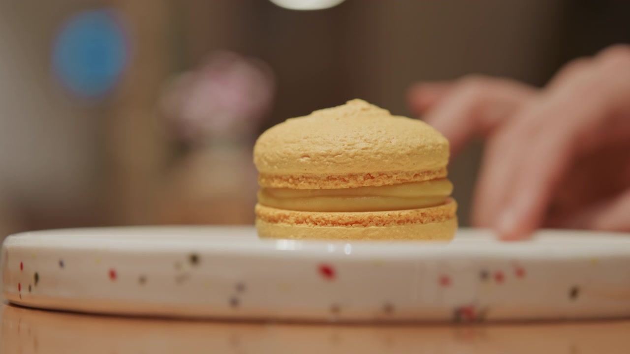 Primo piano di un macaron giallo che ruota su un piatto bianco con macchioline rosse e nere. Una mano appare occasionalmente per regolare la rotazione, rivelando una luce soffusa in un ambiente interno con sfondo sfocato.