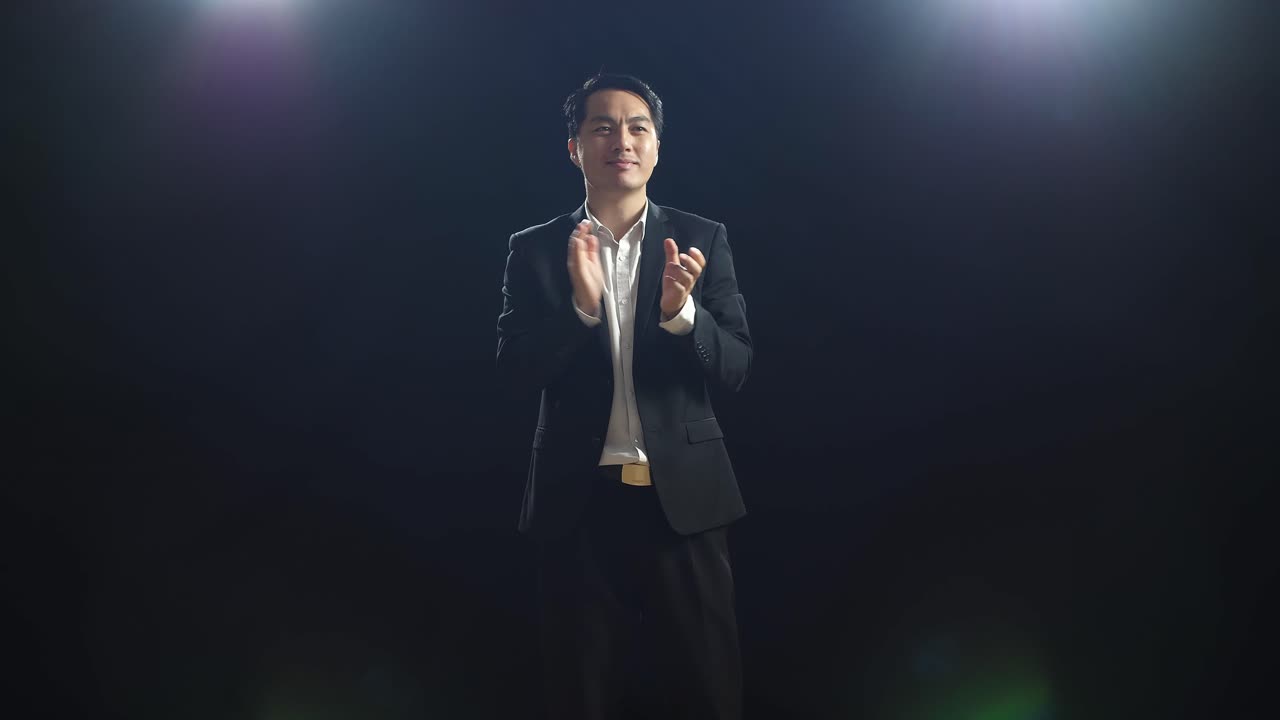 블랙 스크린 스튜디오에 서 있는 동안 손을 박고 주변을 둘러보는 미소 짓는 아시아 스피커 남자