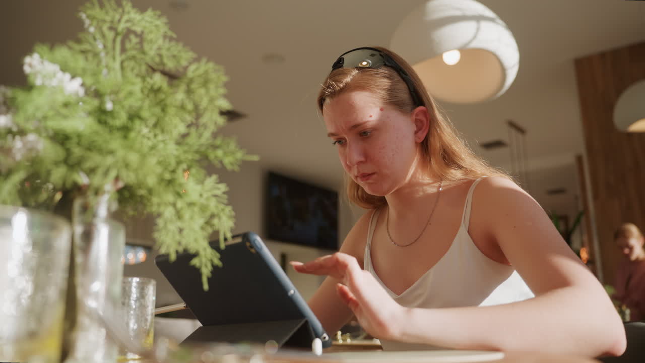 White woman focused typing on tablet, intense expression with pendant necklace, lamp and glass vase in foreground, busy cafe interior with warm evening light, multitasking on email and spreadsheets
