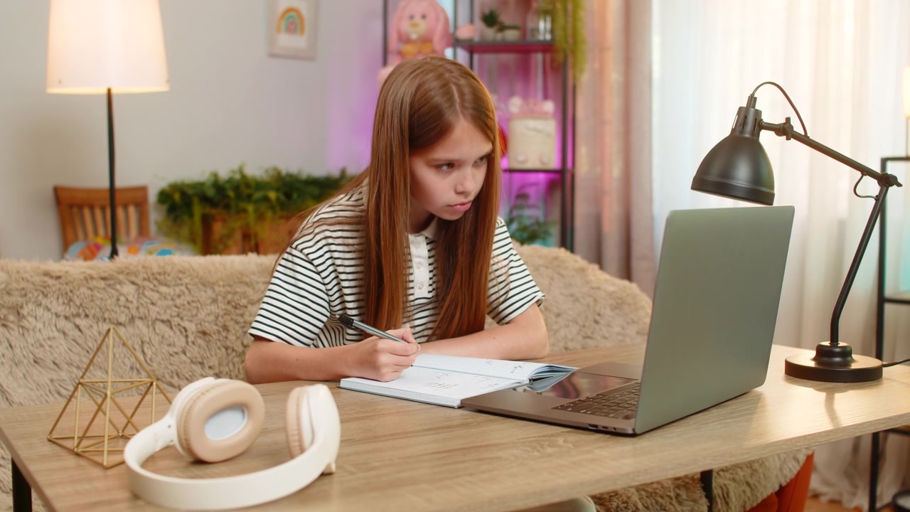 Child girl kid doing online homework on laptop and writing math notes while sitting at home table