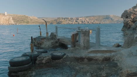 sea water surrounds an old, abandoned boat dock sitting in the foreground with the rocky cliffs of a maltese island in the background