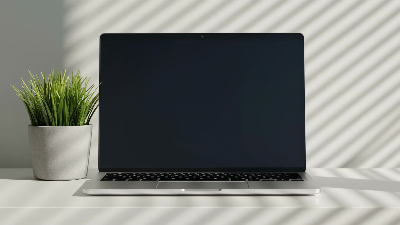 Laptop con schermo scuro posizionato accanto a una pianta verde su un tavolo bianco, mentre la luce solare crea ombre diagonali sulla parete, mostrando una tranquilla disposizione di uno spazio di lavoro interno con toni neutri e una cornice semplice.