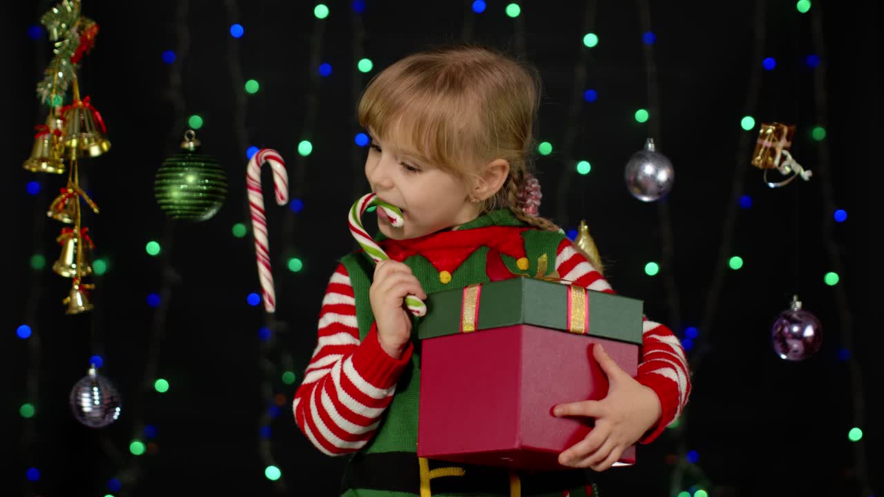 Malá dívka v kostýmu vánočního skřítka, pomocníka Santa Clause, líže lízátko ve tvaru cukrové hole a karamelové sladkosti