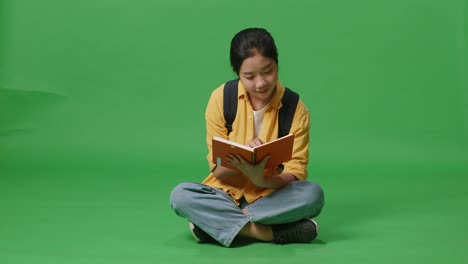 full body of asian woman student with a backpack holding and reading a book while sitting in the green screen background studio