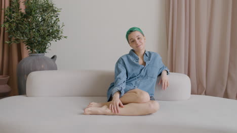 young woman with blue hair and wearing a denim shirt sitting on a big sofa looking at camera